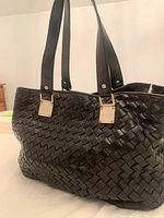 Side view of black leather woven handbag showing double shoulder straps with gold metal plaques stamped with 'Michael Kors'