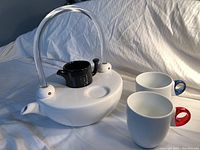 Full view of the white ceramic tea pot with clear acrylic handle, black ceramic teapot insert, and two ceramic mugs with acrylic handles (red and blue).