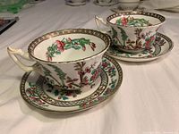 Side view of two floral Coalport tea cups and saucers, showing detailed floral patterns and gold trim on rims and handles.
