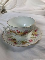 White porcelain tea cup and saucer with floral patterns and gold trim, shown on white fabric background.