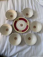 Six white soup bowls arranged in circle with single saucer in center. Bowls have cranberry and floral pattern inside with matching design on saucer.