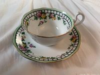 White porcelain tea cup with floral pattern and gold trim on rim and handle, placed inside matching saucer.