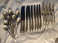 Overall set of silverware pieces including spoons, knives with black blades, and forks arranged in groups on a white cloth showing the pattern and number of pieces.