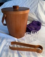 Wooden ice bucket with lid, two small glass tumblers, wooden tongs, and purple cloth bag shown on white fabric background.