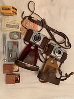 Two vintage cameras, Zeiss Ikon Contina and Silette Pronto, laid out with leather cases, straps, film box, light meter, and accessories on a white surface.