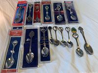 Photo showing 14 vintage collectible souvenir spoons, 6 in original packaging and 8 loose spoons with various designs and decorative tops.