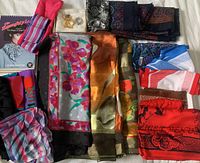 A large assortment of vintage scarves arranged on a white surface, including scarves with floral and geometric patterns in various colors such as red, blue, purple, orange, and green. Scarf clips displayed in a box with an instruction manual is also present.