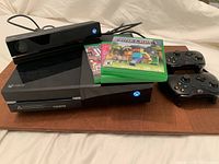 Xbox One console with Kinect sensor, controllers, and three game cases on a wooden board