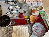 Photo showing assorted serving plates, bowls, decorative platters, glassware, and boxed items on table.