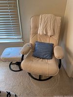 Beige leather chair with cushioned armrests and matching ottoman, both on black circular swivel bases. Cream cable-knit throw blanket draped on chair back and blue pillow on seat.