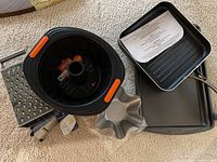 Photo showing black silicone Le Creuset Bundt pan with orange handles, square grill pan, flat baking sheet, two box graters, and small flower shape metal pan on carpet.