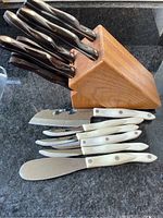 Wooden knife block with five black-handled knives inserted; white-handled knives laid out