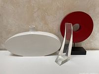 Front view of white oval vase, red round sculpture, and clear lucite sculpture on white table against beige wall