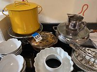 Yellow Dansk pot with lid, Fire King amber casserole dish with lid, white bowls, silverplate pitcher, glass cake stand, silverware forks, and ceramic Bundt pan shown on kitchen stove.