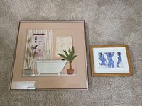 Two framed artworks laid on carpet floor: larger mixed media 'Basically Beige' bathroom scene on left; smaller framed blue watercolor and etching nude signed J Thompson on right.