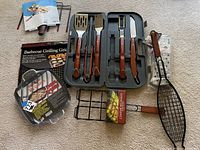 Full lot overview showing grilling tools case, corn cob basket in packaging, barbecue grilling grids and Texas style griddle on carpeted floor.
