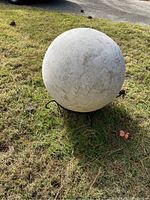 Photo of large round light gray outdoor ball sculpture on grass, resting on metal stand, showing textured spherical surface and outdoor setting.