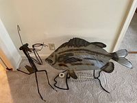 Photo showing metal fish table and metal grasshopper sculpture together on carpeted floor near wall outlet.