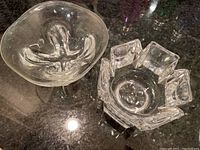 Top view of two clear glass bowls on a dark reflective surface, showing one geometric faceted bowl signed Orrefors and one smooth rounded bowl with an organic shape.