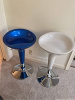 Two adjustable height bar stools, one blue and one white, shown side by side against a wall on carpet flooring.