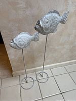Pair of white fish sculptures with speckled black pattern on thin metal stands with circular bases shown against a beige textured wall and tiled floor.