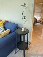 Full view of the round black metal side table with the chrome curved lamp placed on top, positioned beside a blue couch.