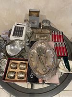 Overview of silver plated serving items on glass table showing napkin rings, shakers, bowls, boxed serving forks and spoons, and serving tray.