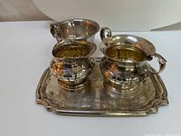 Sterling silver creamer, sugar bowl, small tray and small bowl displayed on a white surface. Items show tarnish and reflective silver finish.