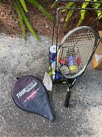 Photo of the Spalding Tour Series tennis racket leaning against a metal holder with several tennis balls inside, placed on concrete outdoors near some foliage. Tennis racket cover with Spalding branding sits on the ground.