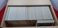 Photo shows a cardboard box filled with many trading cards packed upright. Most cards show backs with no visible front images except a few edges of sports-themed cards with player images and branding.