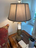 First lamp on wooden side table near a couch, showing beige lampshade with dark vertical trim and ribbed brass base with palm leaf accents.
