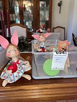 Photo showing one bunny doll seated outside container and other dolls partially visible inside clear plastic container on a wooden surface