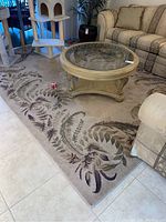 Photo showing the large area rug with leaf design, beige color, and plush texture with part of glass coffee table and sofa visible.