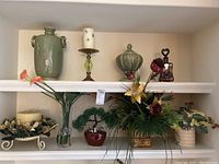 Photo showing shelf with various ceramic vases, glass vases, faux plants with planters, and candles arranged closely together.