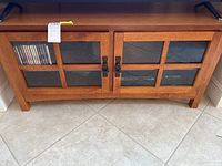 Front view of wooden entertainment console showing glass paneled double doors with six panes each, metal handles, and visible storage inside, sitting on tile floor.