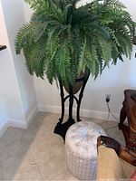 Faux fern plant on composite plant stand next to patterned footstool on tiled floor, corner of a room.