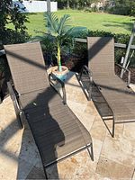 Two brown woven fabric poolside lounge chairs with dark metal frames and armrests, placed on stone tile flooring near garden plants, accompanied by an artificial palm in a ceramic planter.