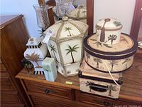 Photo shows 7 decor items grouped on a wooden surface: large hexagonal urn with palm design, white urn with gold leaf design (damaged), palm tree hat boxes, glass figurine, candle holder, and candle.
