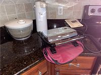 Photo of salad spinner, coffee dispenser, and Hamilton Beach panini maker on kitchen counter with instruction manual and red oven mitt beneath panini maker.