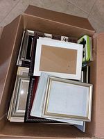 Box filled with assorted picture frames in a variety of finishes and sizes stacked inside, showing mixture of silver, black, and white frames.