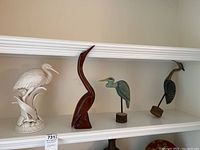 Four bird sculptures displayed on white shelf including a white ceramic bird, tall reddish-brown wood bird, and two smaller wood birds painted green and brown.