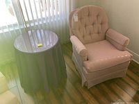 Photo showing the pink upholstered swivel chair and the round table with cloth and glass top placed next to a window on wood flooring.