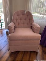 Front view of pink upholstered swivel chair showing tufted backrest, armrests, and skirted base on wooden floor near window.