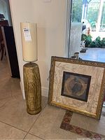 Tall candle with carved decorative base and large off-white candle on top, standing upright next to the framed wall art leaning against the wall.