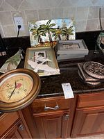 Countertop displays various framed and decorative wall art including one palm tree panel and bird-themed framed print, plus a round clock with floral face and wood frame and ornate metal cheese server with spreader.
