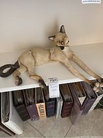 Overall view of the beige ceramic cat sculpture lying on a shelf with elongated legs and curled tail.