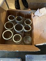 Box containing assorted used glass canning jars upright with metal rings around the openings visible. Cardboard box is worn and open.
