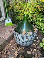 Green glass carboy sitting in a blue-gray wicker basket outdoors, surrounded by fallen leaves and plants.