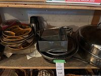 Multiple kitchenware items on a wooden shelf including metal pans, ceramic escargot dishes, square molds, and floral ceramic serving plates.