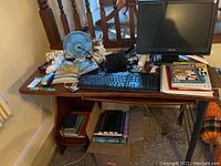 Wide view of wooden desk with monitor, keyboard, blue desk fan, pens, notebooks, and books underneath.